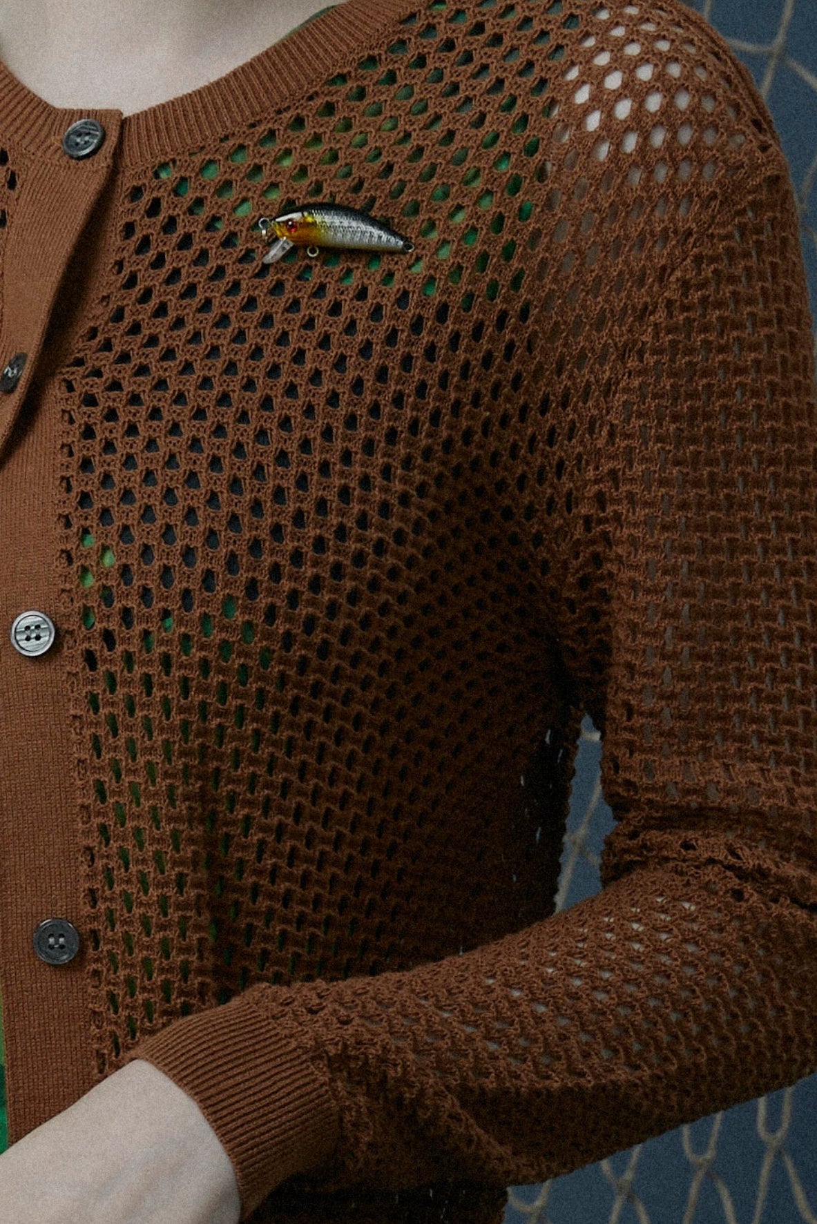 Woman wearing a brown knitted cardigan standing against a blue background with fishing nets.