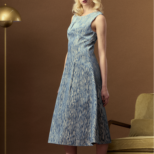 A blonde woman models a sleeveless, knee-length blue and white patterned dress in a styled indoor setting with a gold floor lamp and a green armchair against a brown wall.