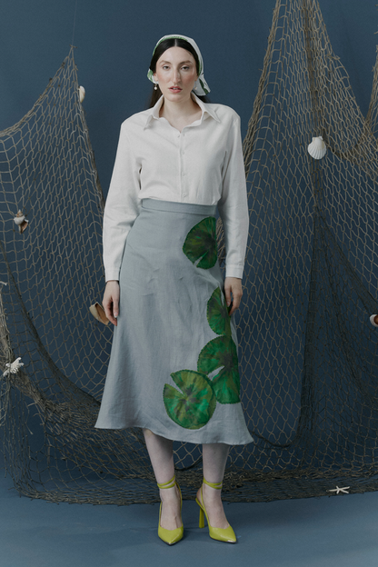 Woman in a white blouse and blueish grey  skirt with hand drawn lily pads applique'd on the left hip to the hem standing in front of fishing nets.