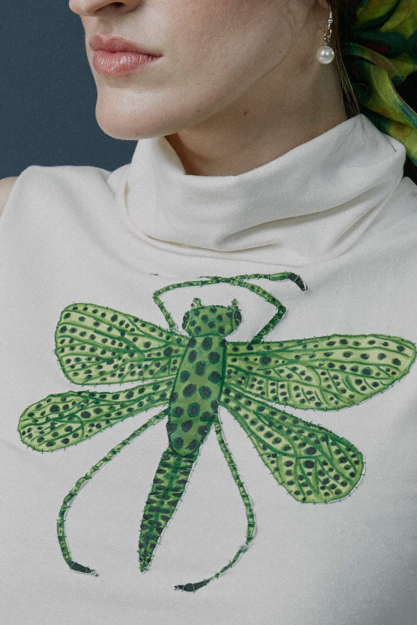 Woman wearing a sleeveless top with a green dragonfly design and a patterned headscarf against a textured wall.